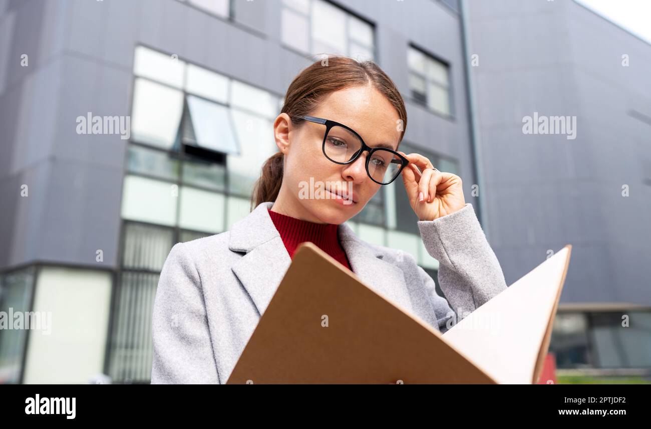 Facility manager woman wearing glasses reading project documentation outdoors standing near near office building. Stock Photo