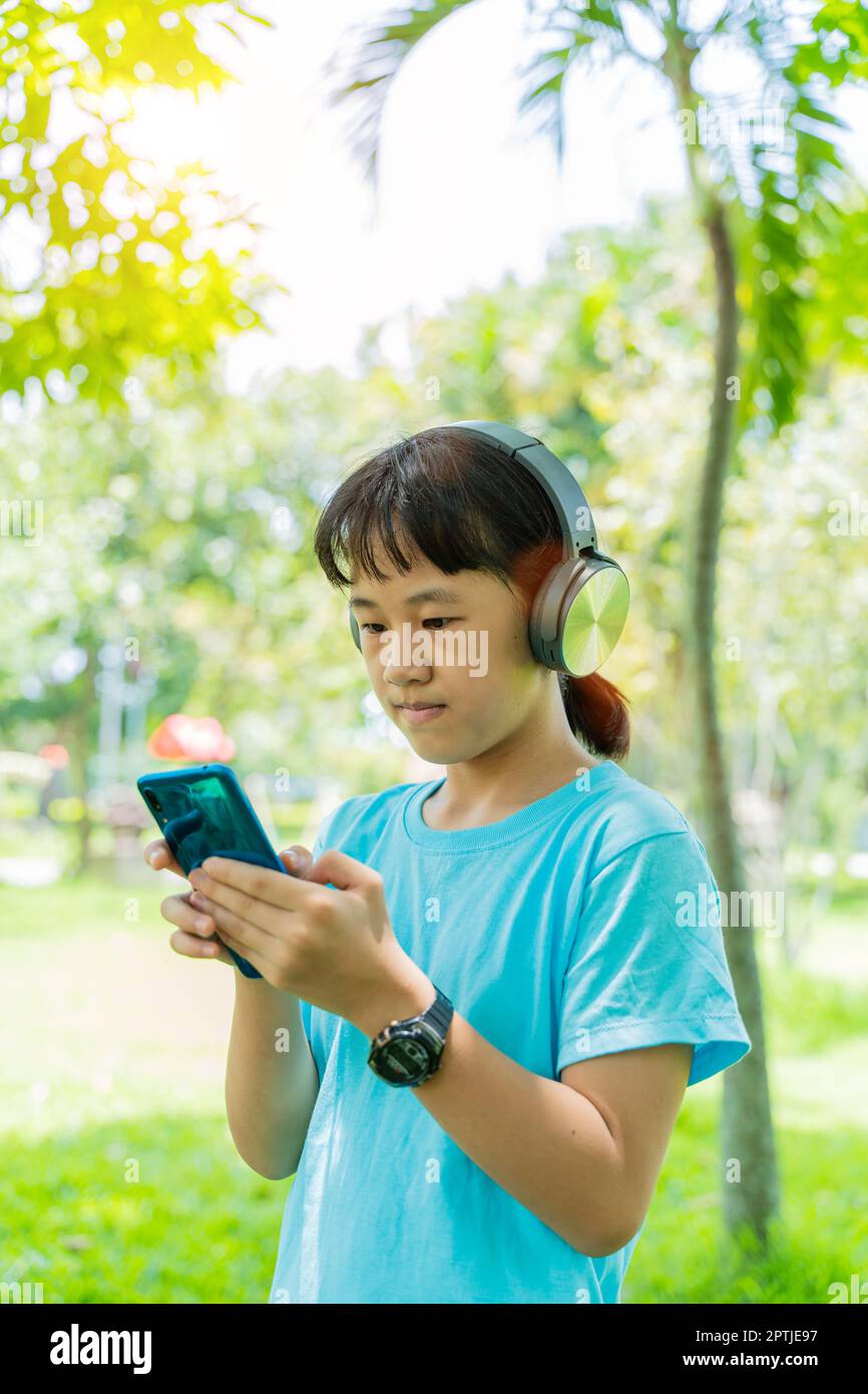 Pretty school girl with smartphone and listens to music with modern headphones wireless in park outdoors. Stock Photo