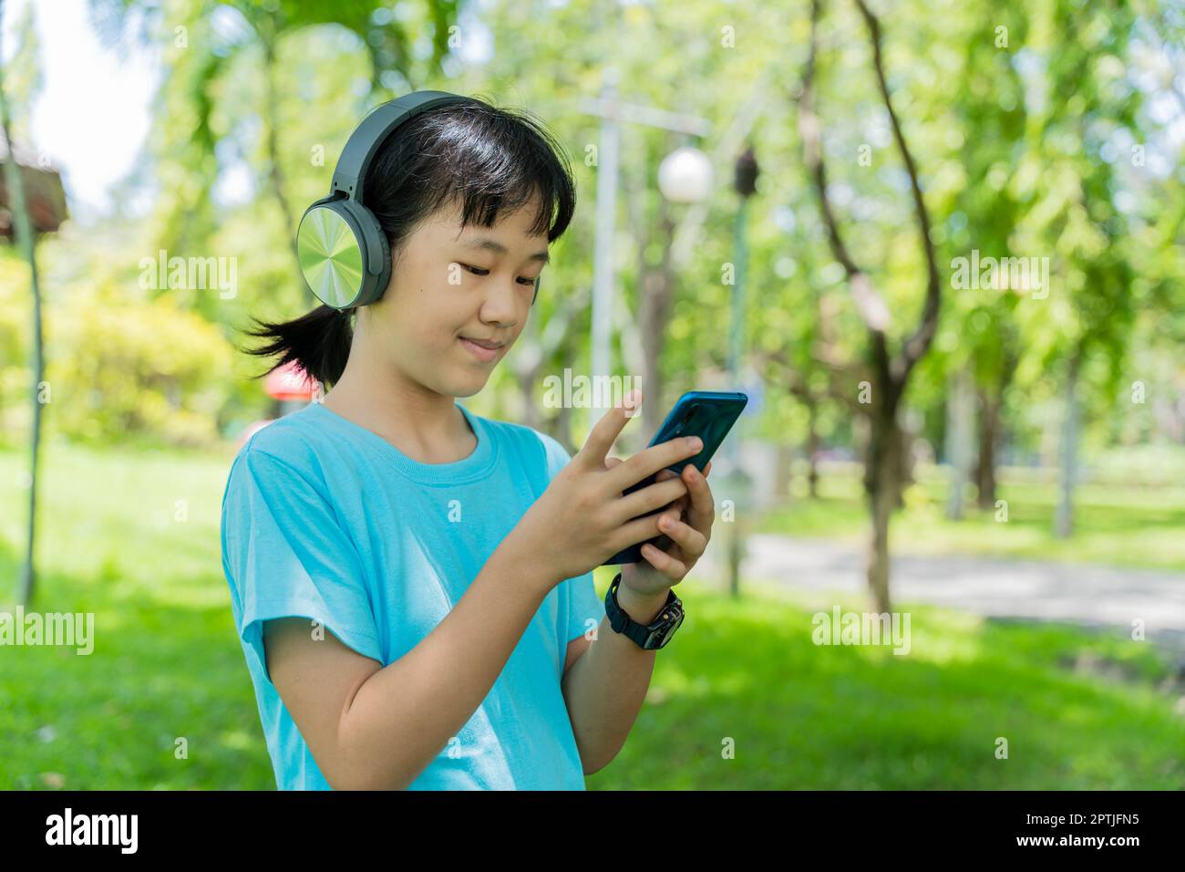 Pretty school girl with smartphone and listens to music with modern headphones wireless in park outdoors. Stock Photo