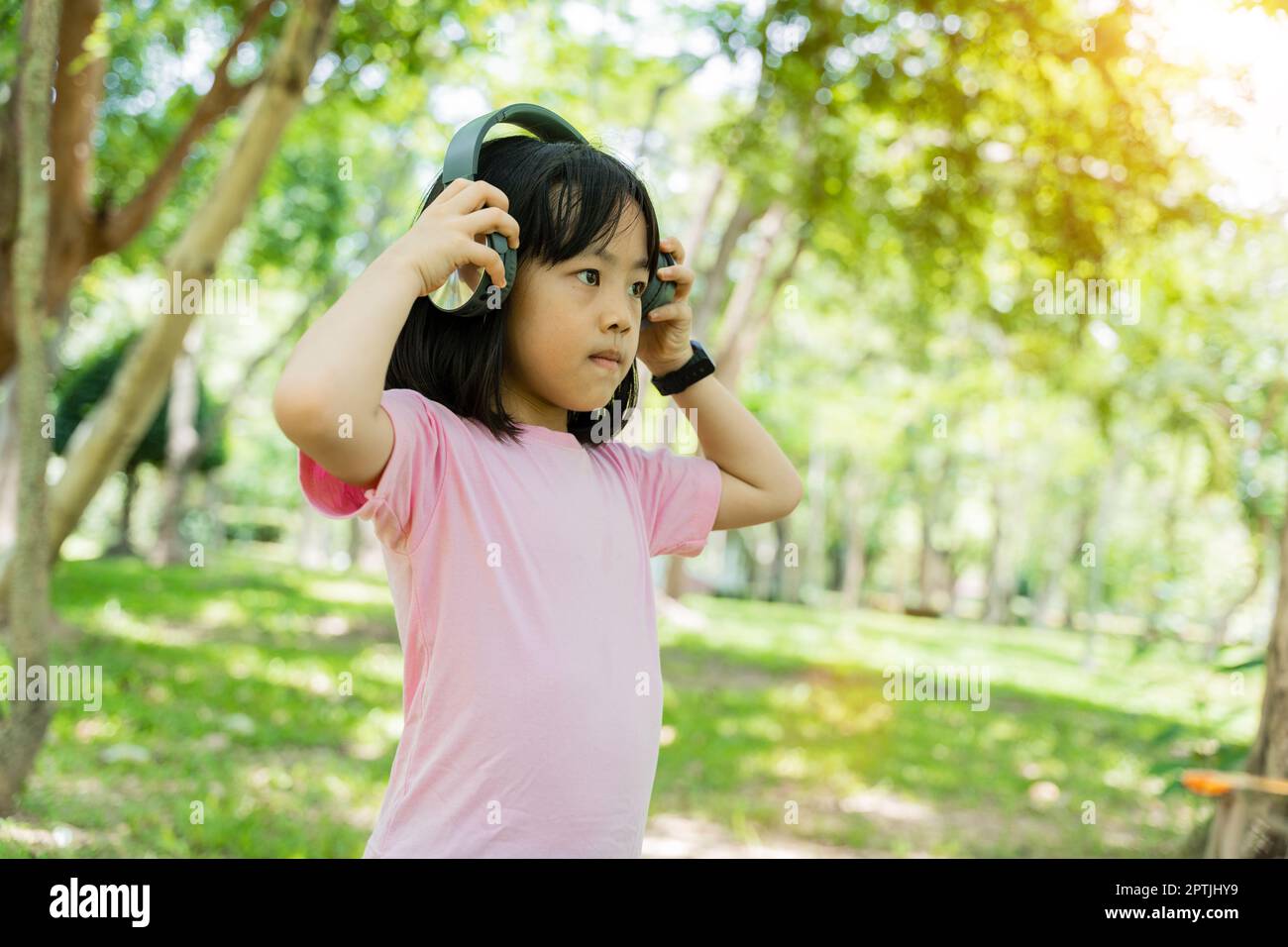 Portrait of child girl listens to music with modern with headphones wireless in green park outdoor. Stock Photo