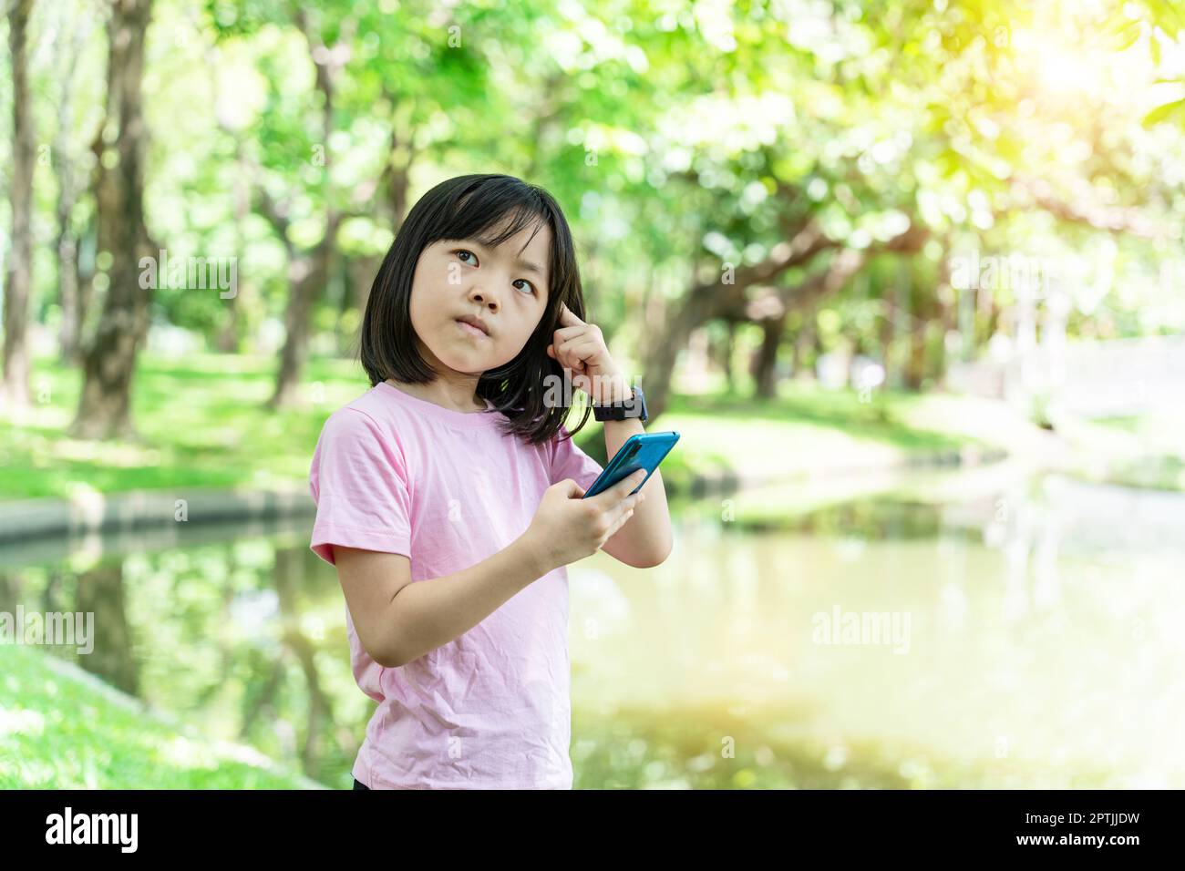 Beautiful asian child girl using mobile smartphone. Little girl is playing in digital game in the green park. Stock Photo