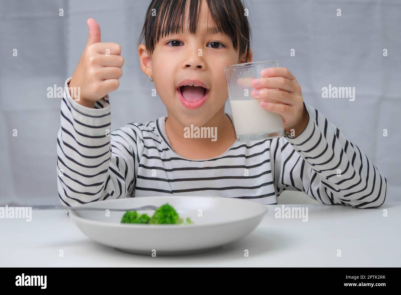 Cute Asian girl drinking a glass of milk in the morning before going to school. Little girl eats healthy vegetables and milk for her meals. Healthy fo Stock Photo