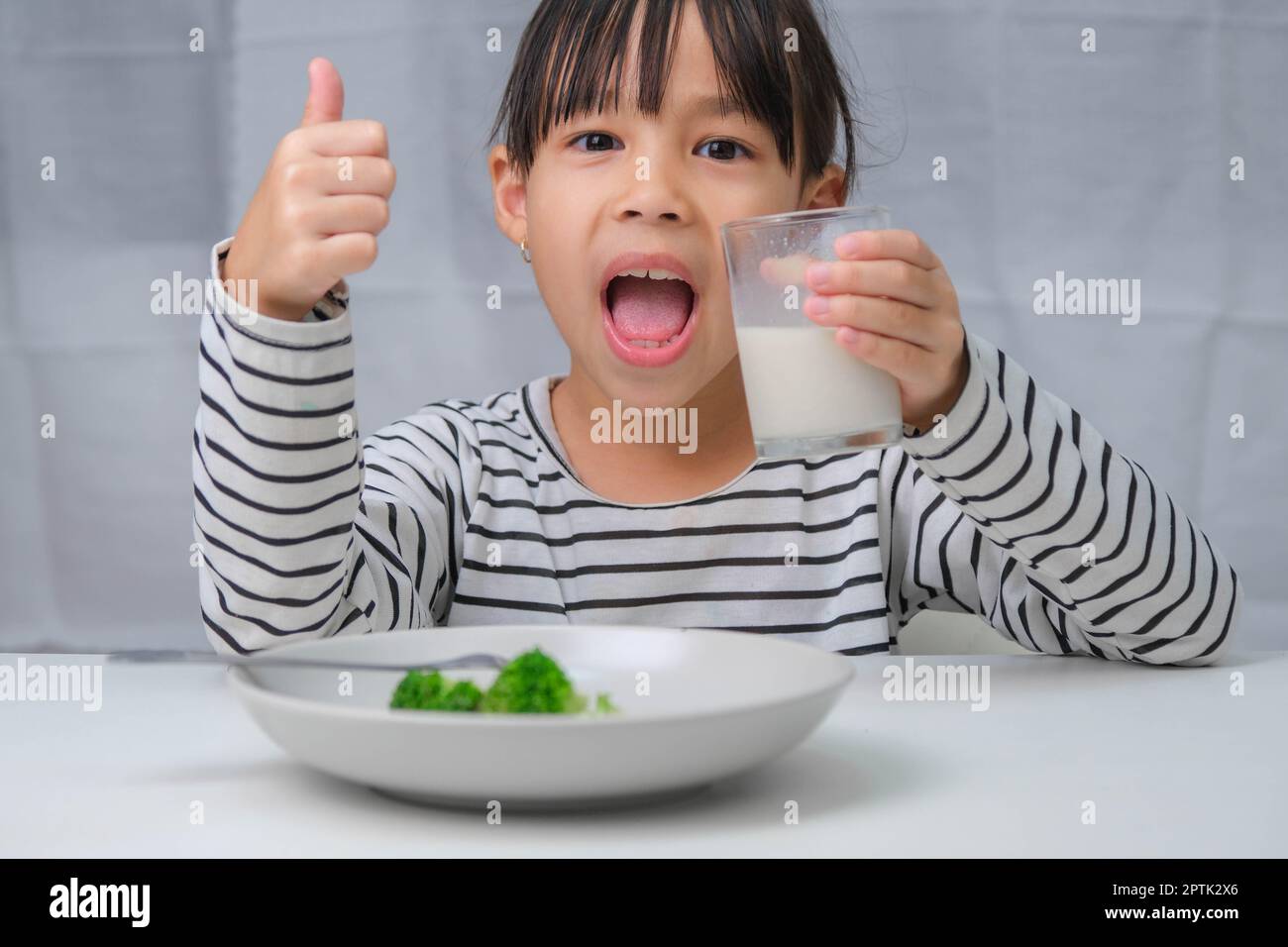 Cute Asian girl drinking a glass of milk in the morning before going to school. Little girl eats healthy vegetables and milk for her meals. Healthy fo Stock Photo