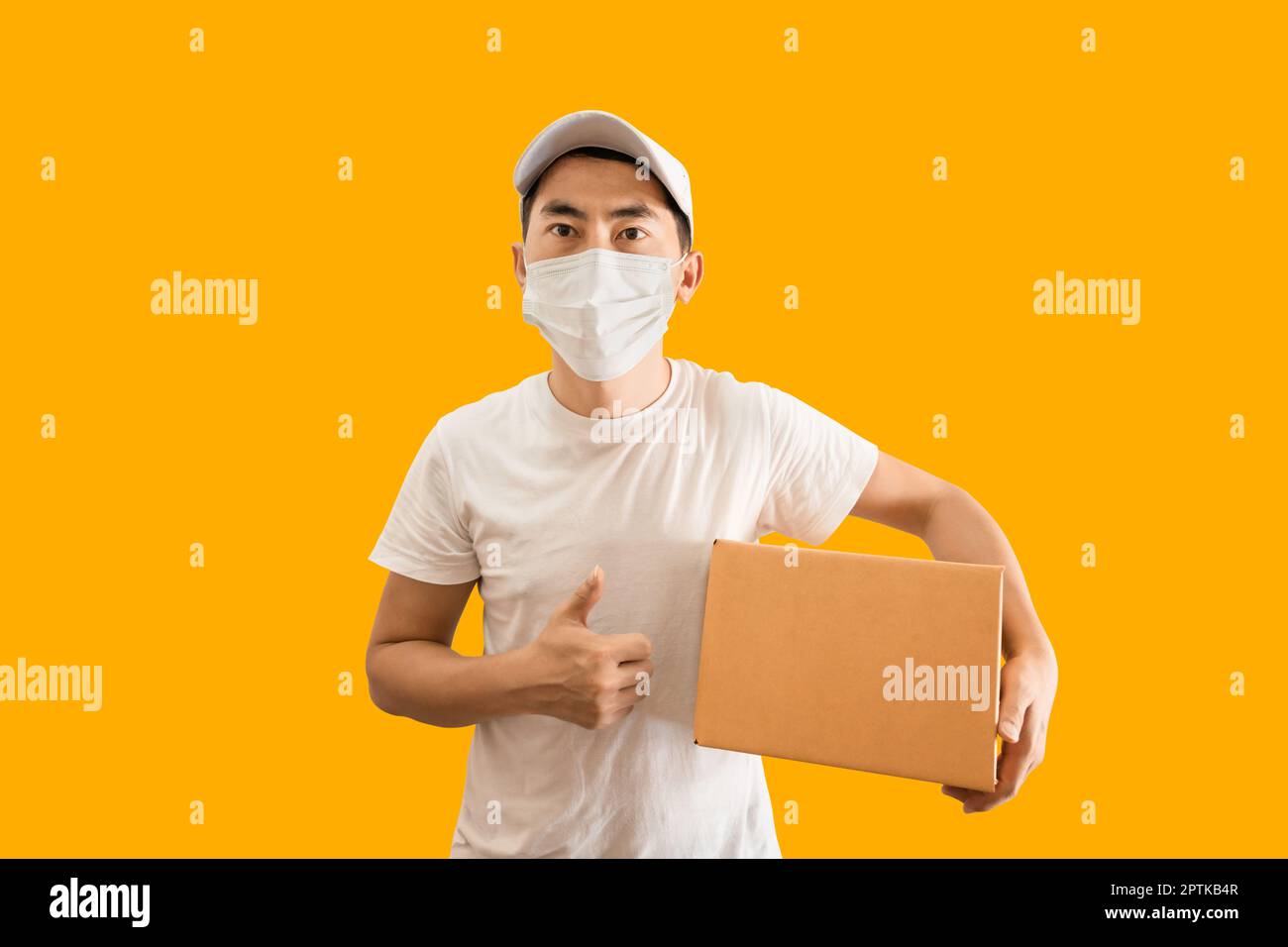 Young Asian delivery man wearing cap and white blank t-shirt holding parcel post box isolated on yellow background. express delivery service concept. Stock Photo
