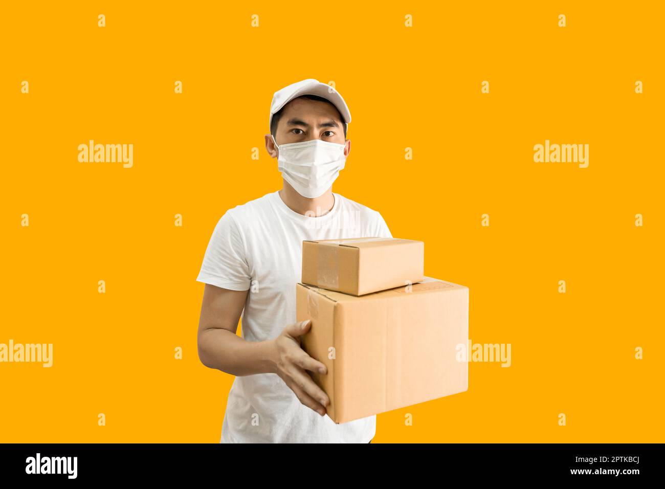 Young Asian delivery man wearing cap and white blank t-shirt holding parcel post box isolated on yellow background. express delivery service concept. Stock Photo