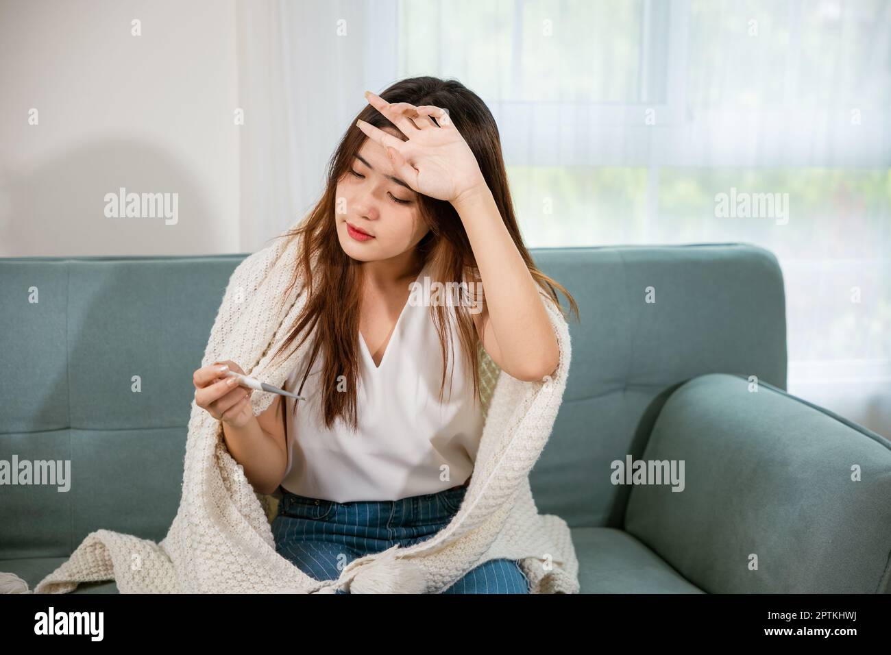 Sick Asian young woman sit under blanket on sofa she sneeze checking temp with digital