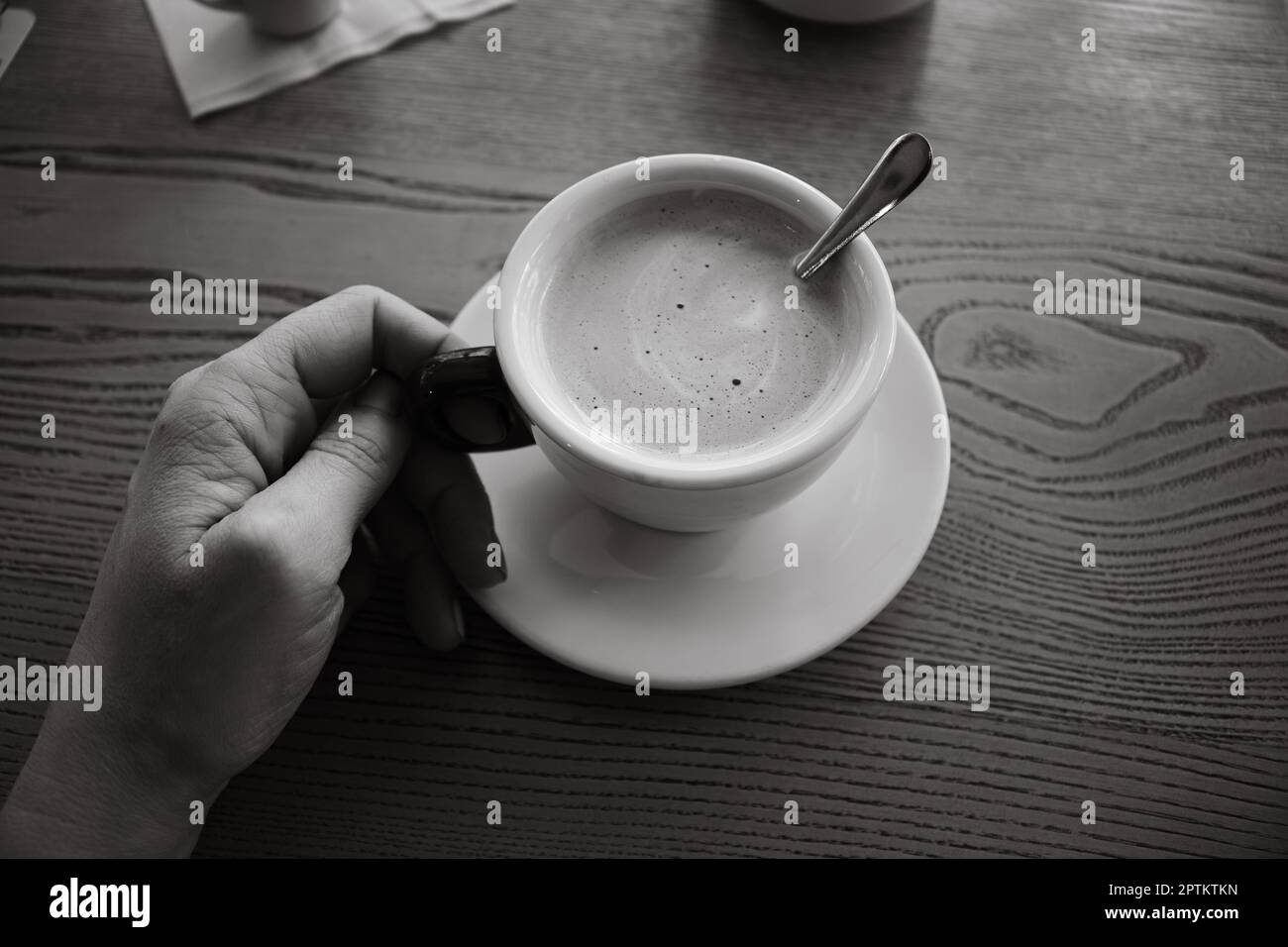 Woman hand holding coffee cup, black and white. Coffee with milk and sugar on table, monochrome. Morning drinks. Girl with cappuccino cup on table. Stock Photo