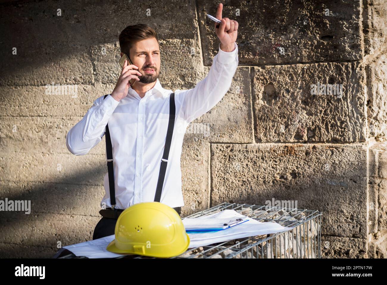 Male architect talking on mobile phone with blueprint at construction site, Munich, Bavaria, Germany Stock Photo