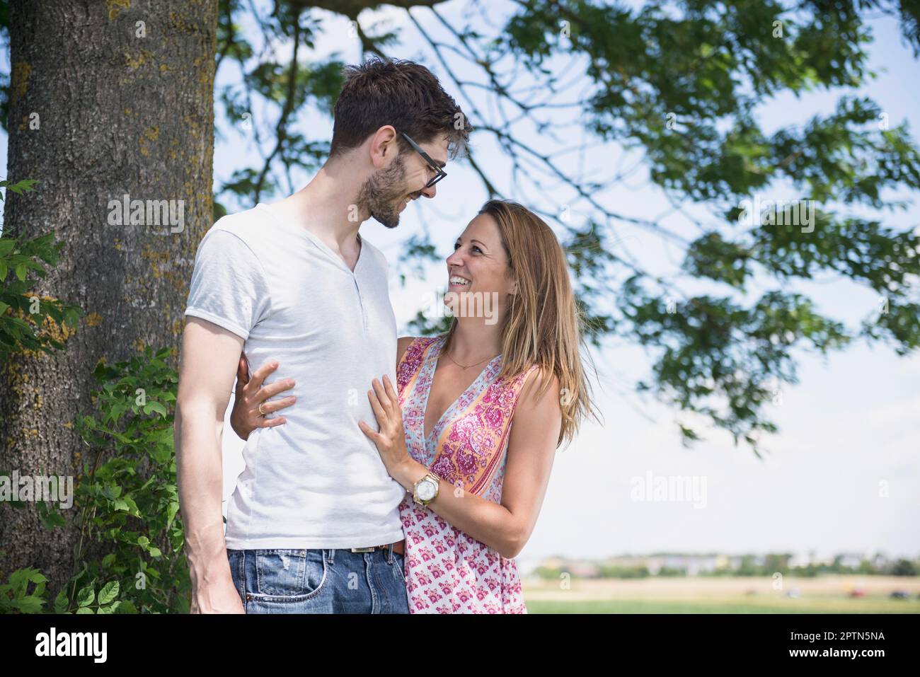 Mid adult couple smiling at each other in front of tree in the countryside, Bavaria, Germany Stock Photo