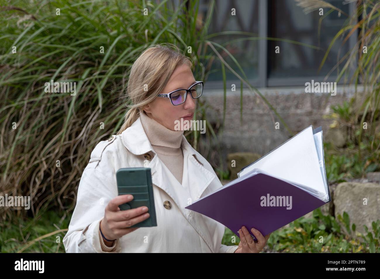Beautiful matur woman stands near glass building with folder and phone Stock Photo