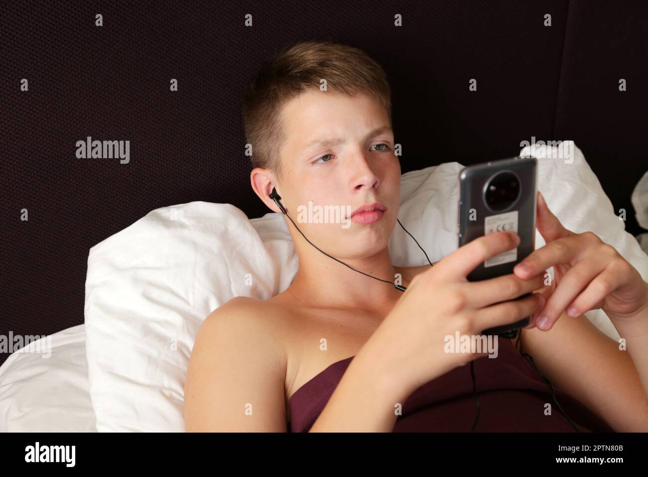 A teenager lies in bed with headphones and a smartphone in his h Stock Photo