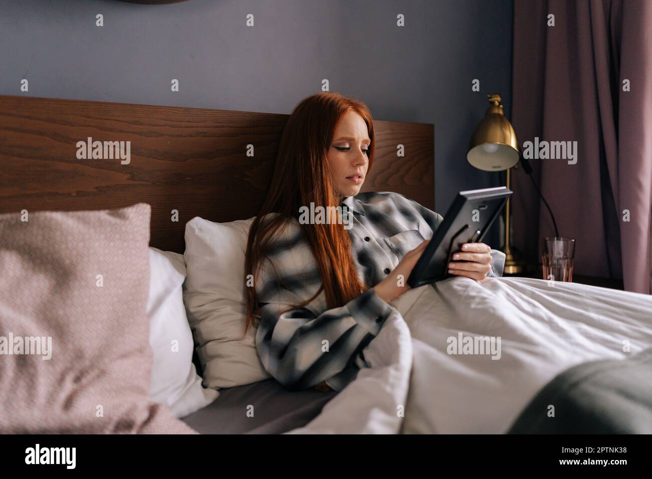 Sad young woman lying on bed under blanket holding photo in frame in