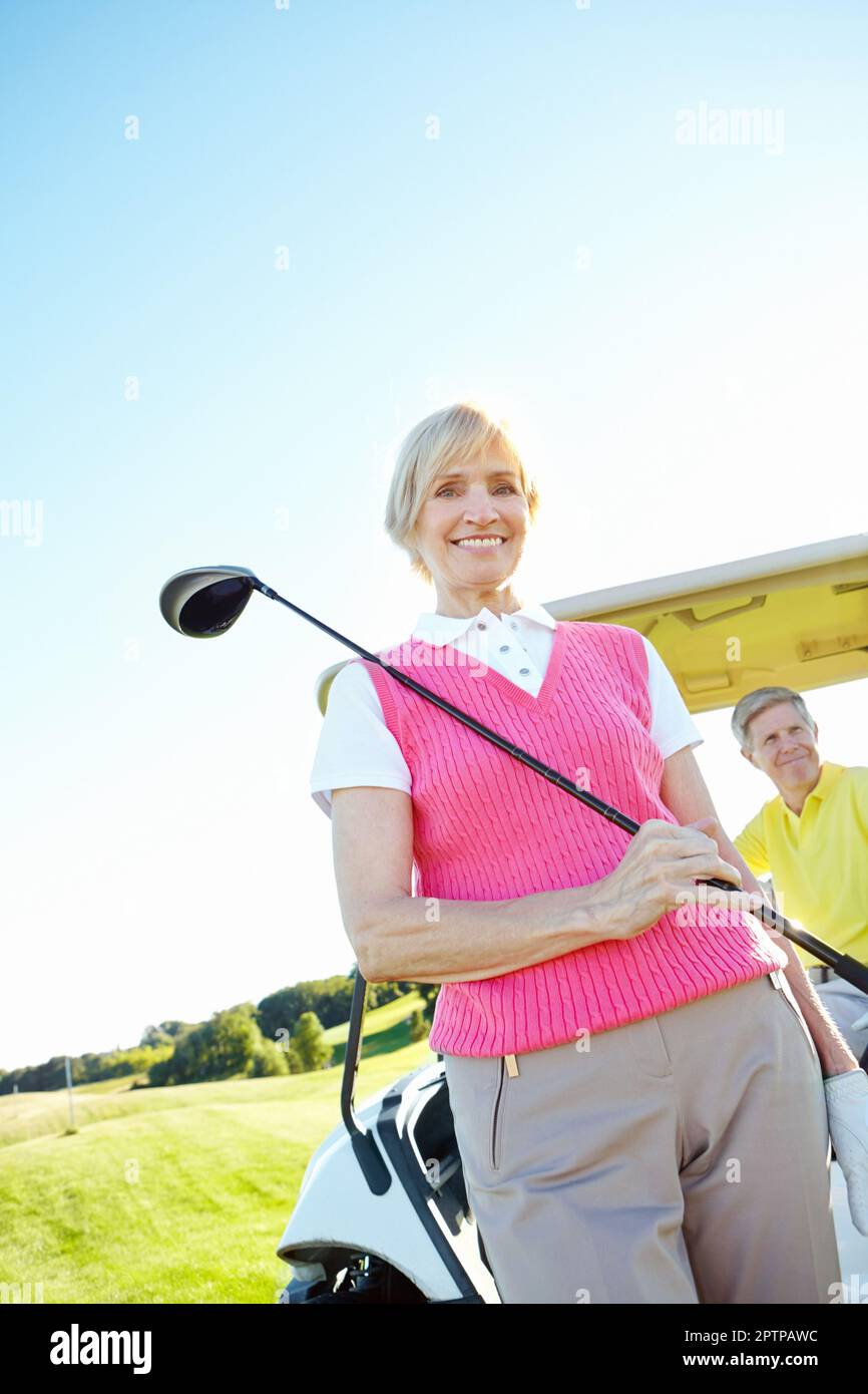 Peaceful pastime. Low angle shot of an attractive older female golfer standing in front of a golf cart with her golfing buddy behind the wheel. Stock Photo