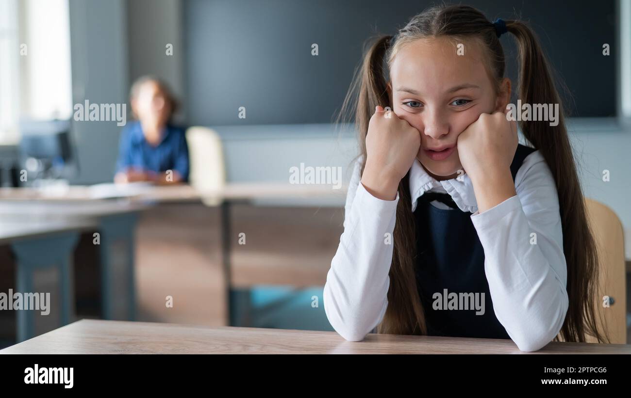 Little caucasian girl is bored at the lesson at school. The schoolgirl is sitting at her desk and the teacher is sitting in the background. Stock Photo