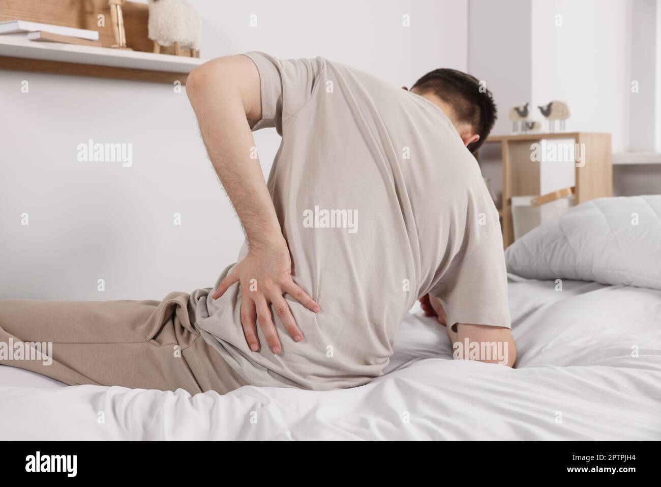 Man suffering from back pain while sitting on bed in room. Symptom of scoliosis Stock Photo