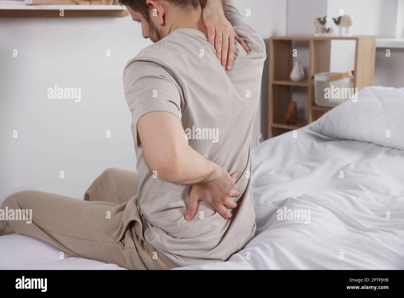 Man suffering from back pain while sitting on bed in room. Symptom of scoliosis Stock Photo