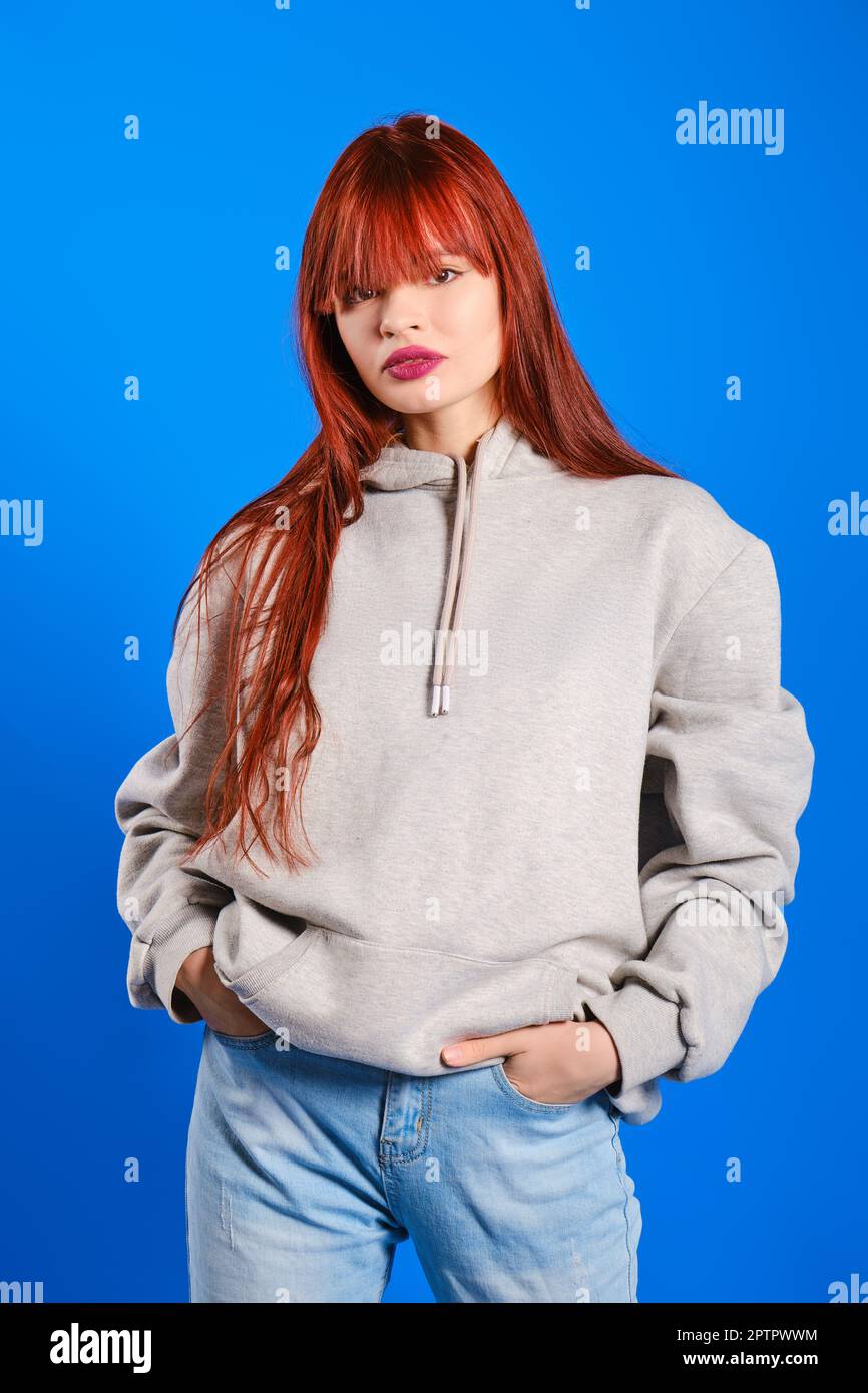 Studio portrait of young woman with red hair in grey hoodie with hands in pockets of her jeans on blue background Stock Photo
