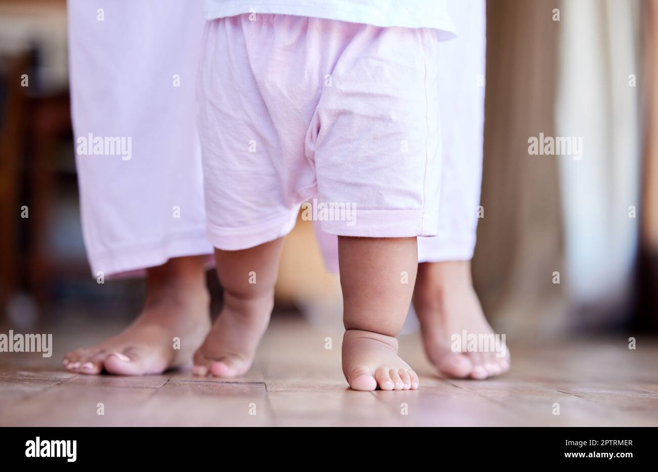 Little baby girl walking with the help of her parent. Closeup of the feet of a small child walking at home with her mother behind her. Mother helping Stock Photo