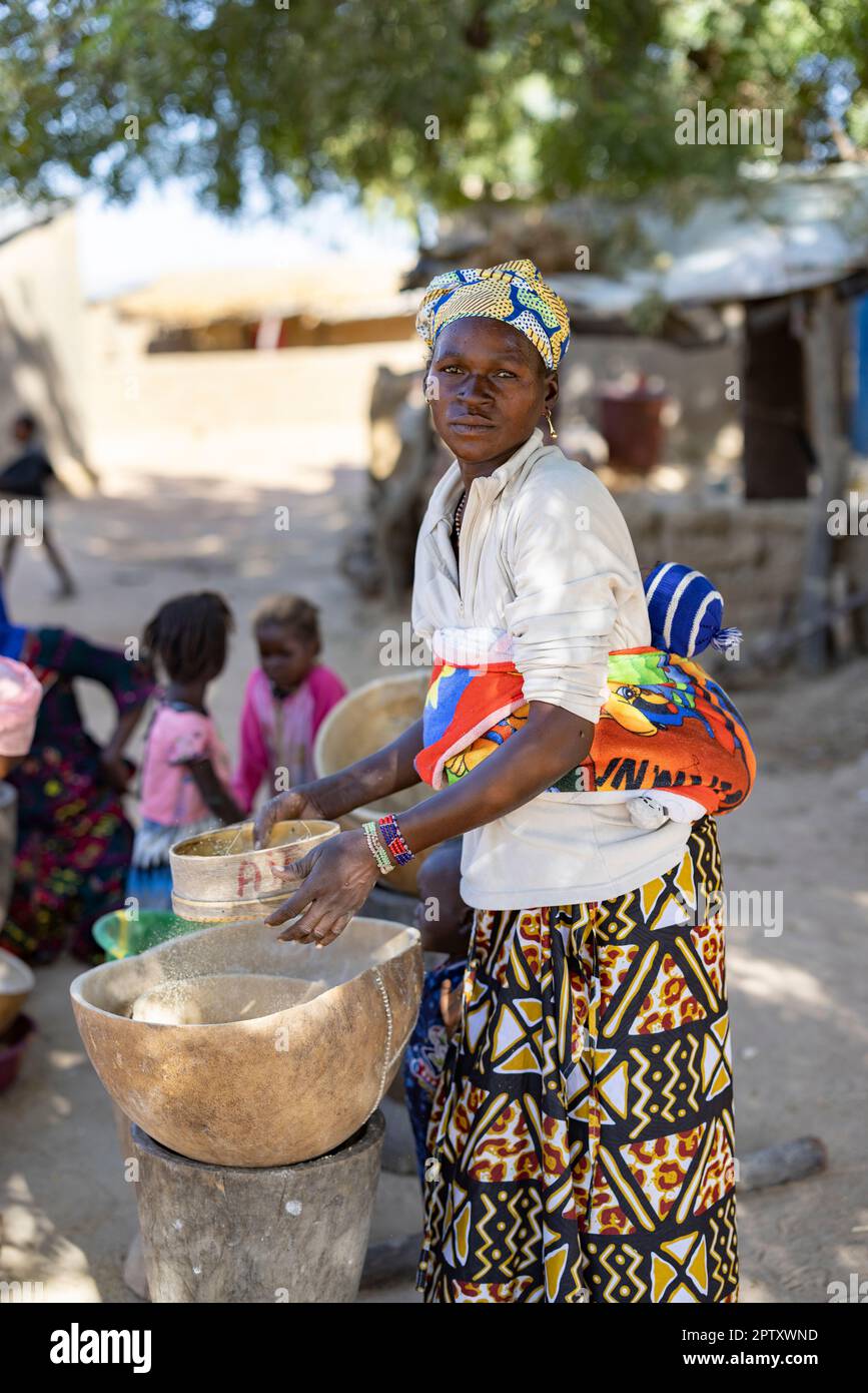 An African woman carrying her baby on her back uses a sieve and calabash to refine her millet grain flour in Segou Region, Mali, West Africa. 2022 Mali drought and hunger crisis. Stock Photo
