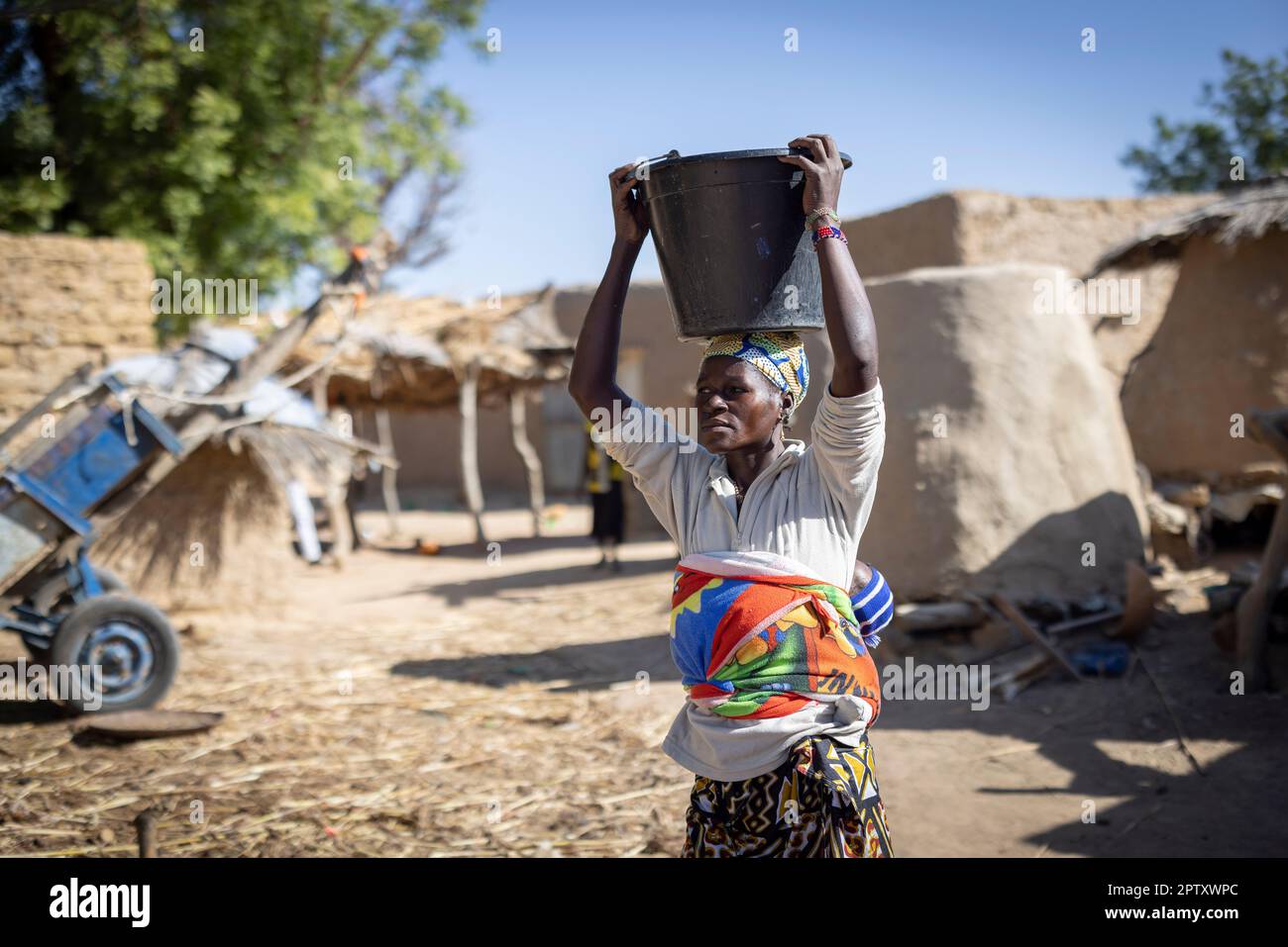 An African woman carries a bucket of water on her head and her baby on her back in Segou Region, Mali, West Africa. 2022 Mali drought and hunger crisis. Stock Photo