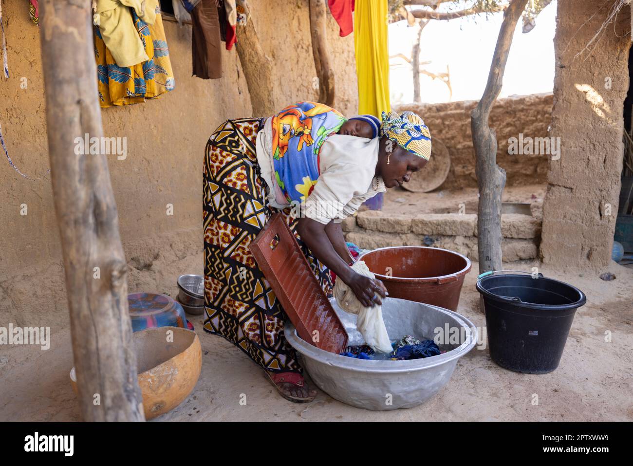 A mother washes clothes at her home while carrying her newborn baby on her back in Segou Region, Mali, West Africa. 2022 Mali drought and hunger crisis. Stock Photo