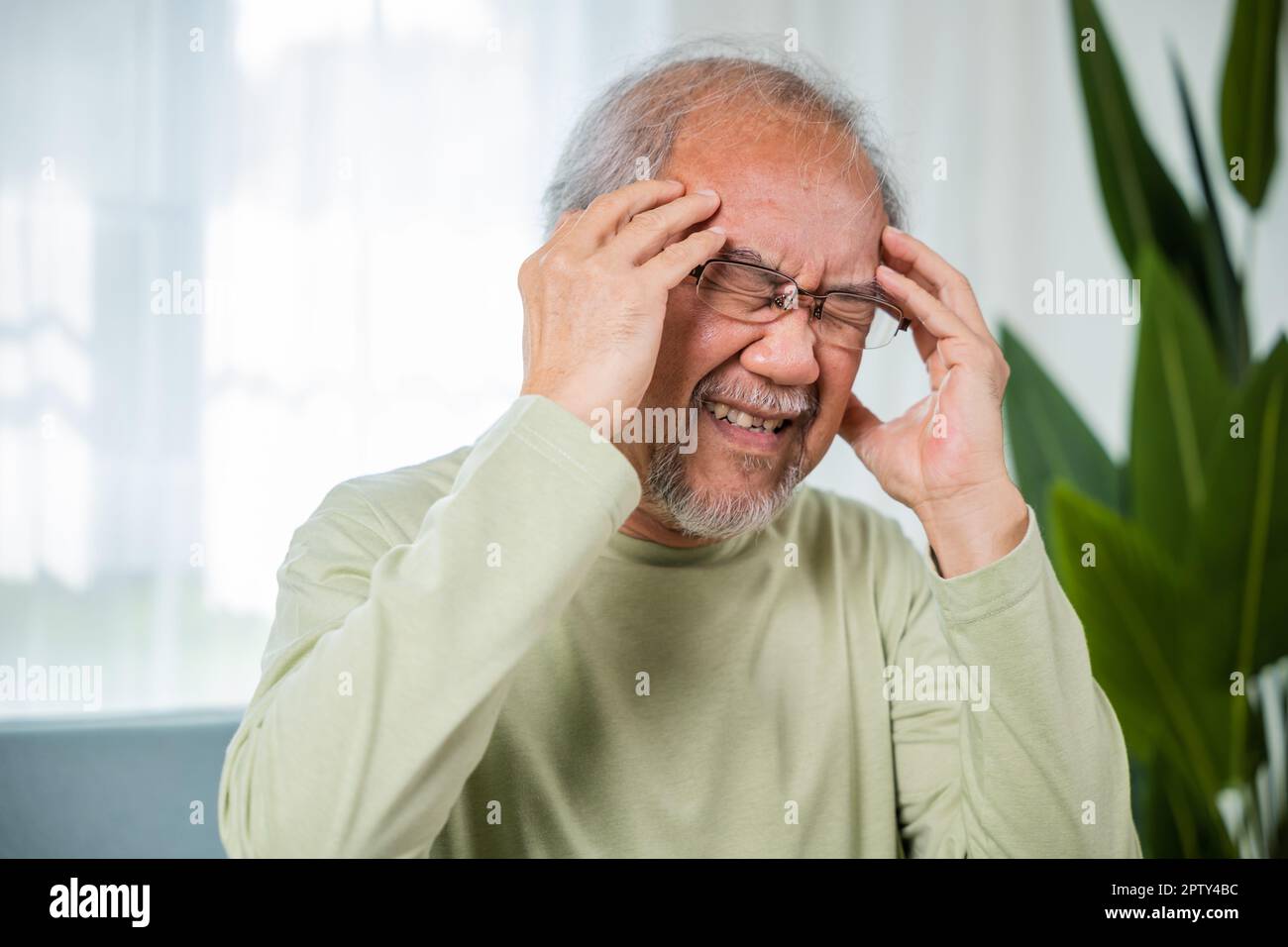 Headache. Close up of elderly holds head with hand suffering from migraine headache, Sad Asian senior man sitting on sofa feeling hurt and lonely at h Stock Photo