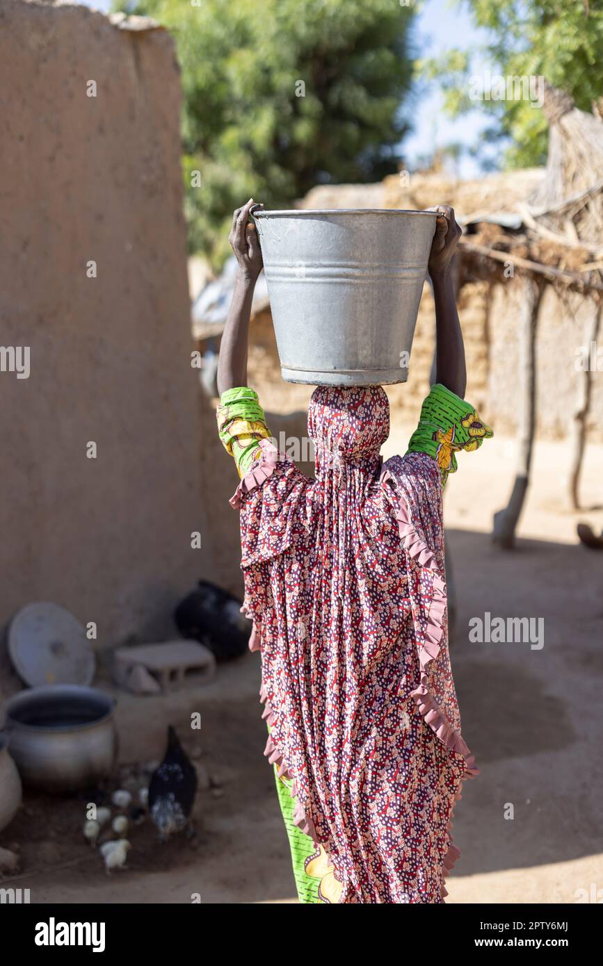 A woman in traditional Africaln dress carries a bucket of water on her head in Segou Region, Mali, West Africa. 2022 Mali drought and hunger crisis. Stock Photo