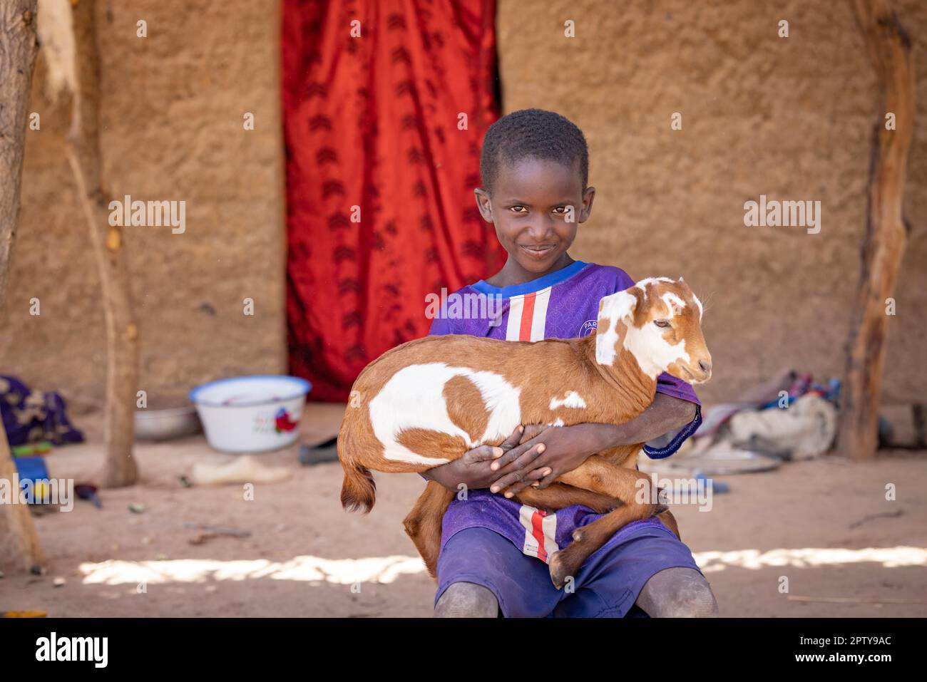 A boy sits outside his mud house holding a goat in Segou Region, Mali, West Africa. 2022 Mali drought and hunger crisis. Stock Photo