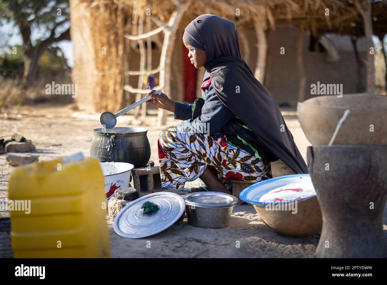 A young Fulani refugee woman, displaced from conflict in the North, boils food over an open fire in Segou Region, Mali, West Africa. 2022 Mali drought and hunger crisis. Stock Photo
