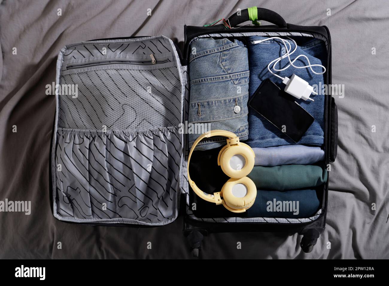 carry-on suitcase with blue clothes and large headphones, charger and cell phone on the bed Stock Photo