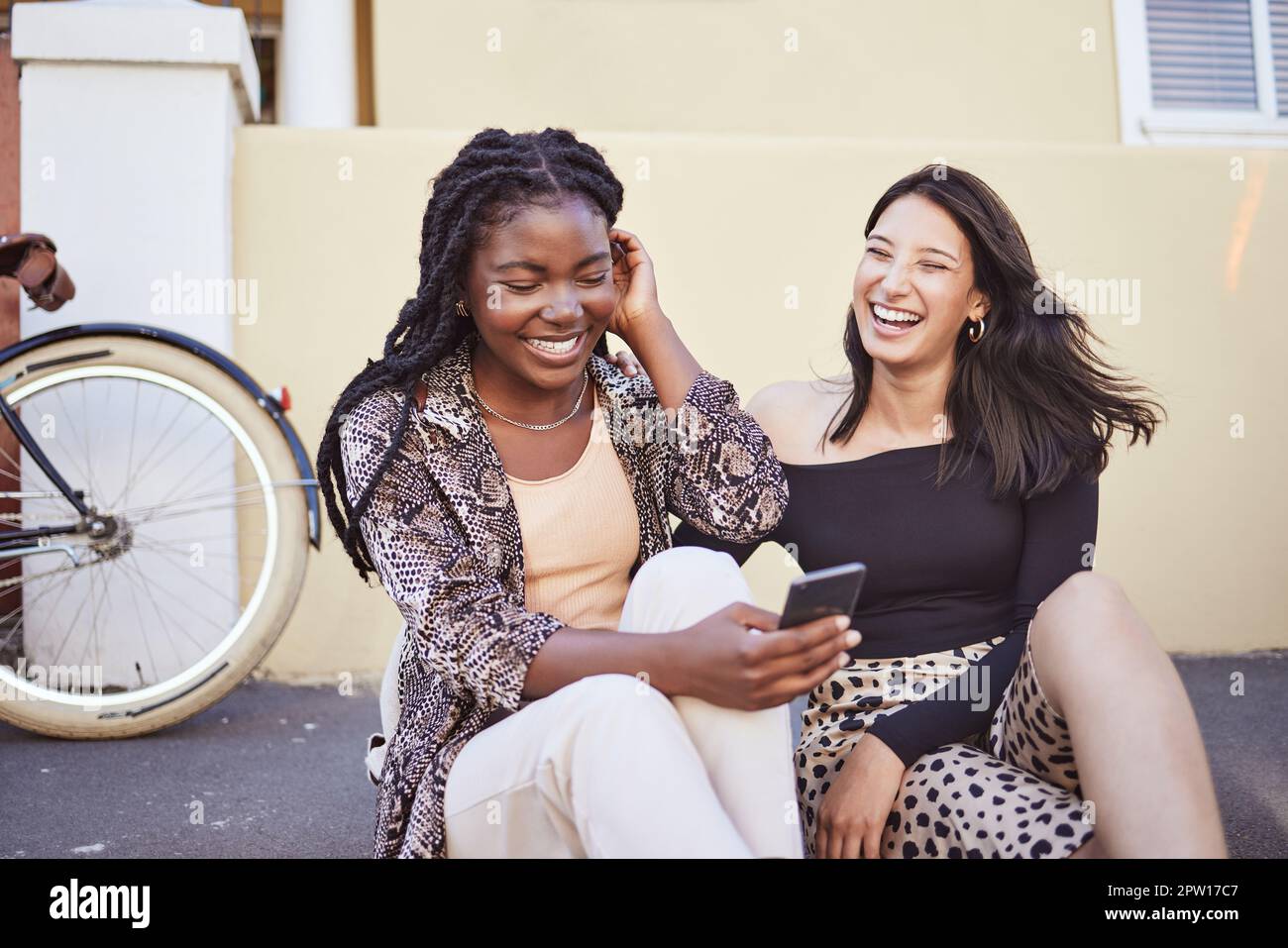 Two multiethnic female friends sitting on sidewalk and laughing while browsing social media. Best friends checking smartphone content while sitting on Stock Photo