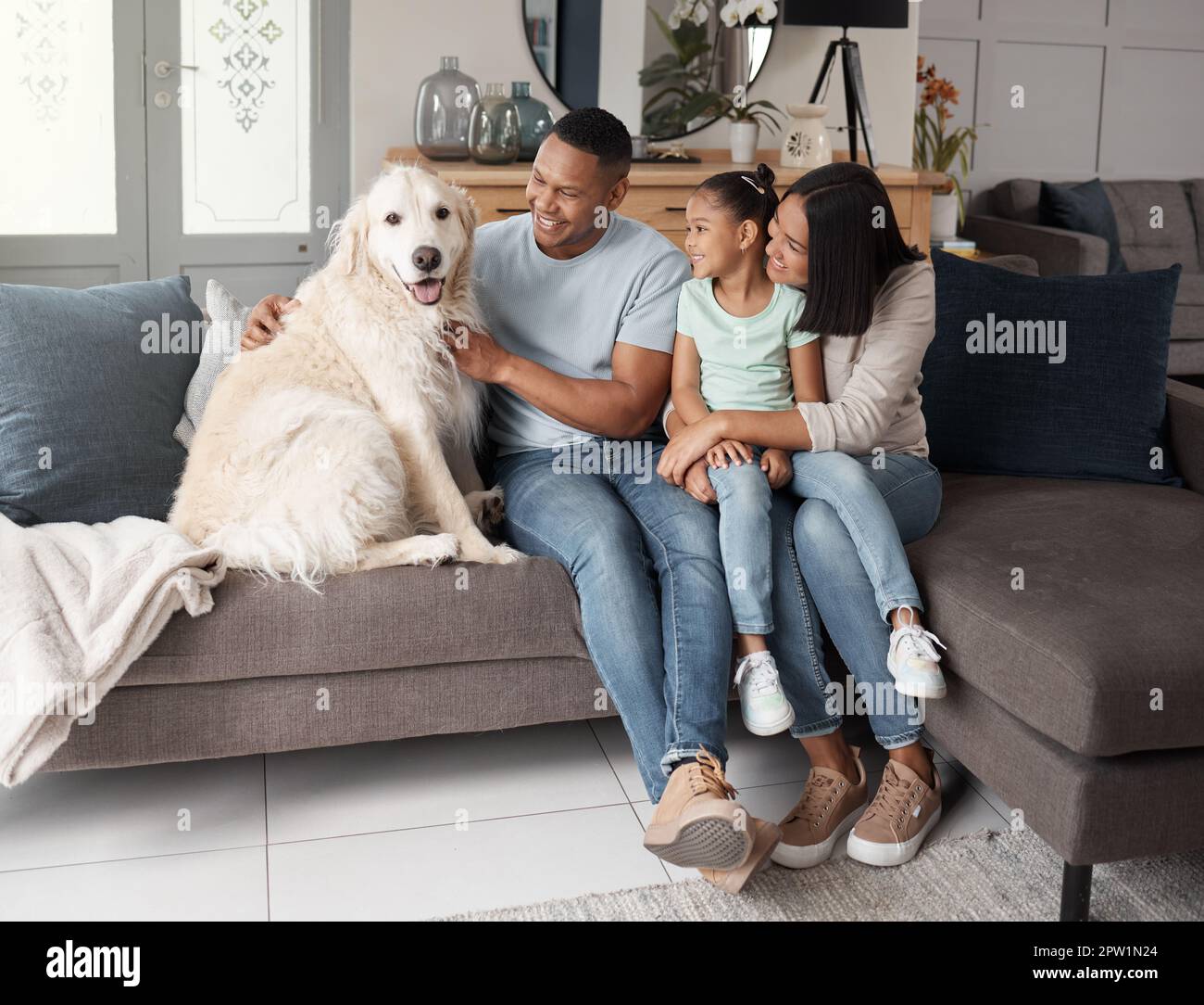 A happy mixed race family of three relaxing on the sofa with their dog. Loving black family being affectionate with a foster animal. Young couple bond Stock Photo