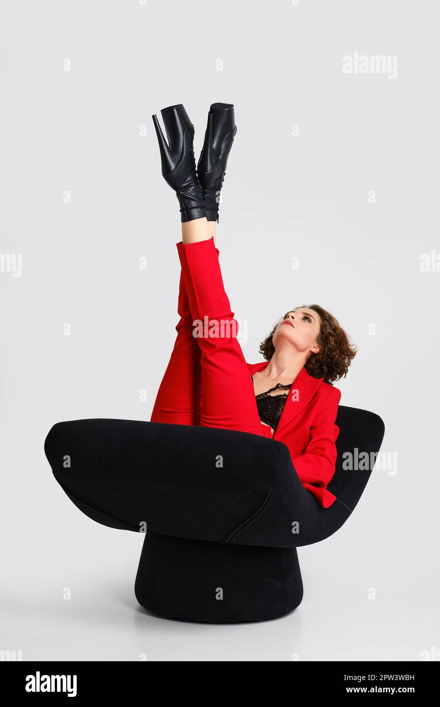 Young woman in red pantsui and pole dance boots lifted crossed legs up in a soft armchair Stock Photo