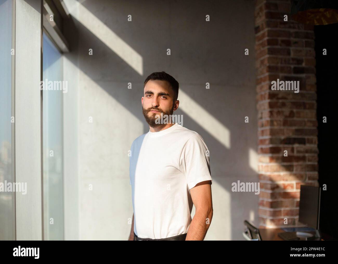 Bearded man in white t shirt looking at camera while standing near window in sunlit loft workplace Stock Photo