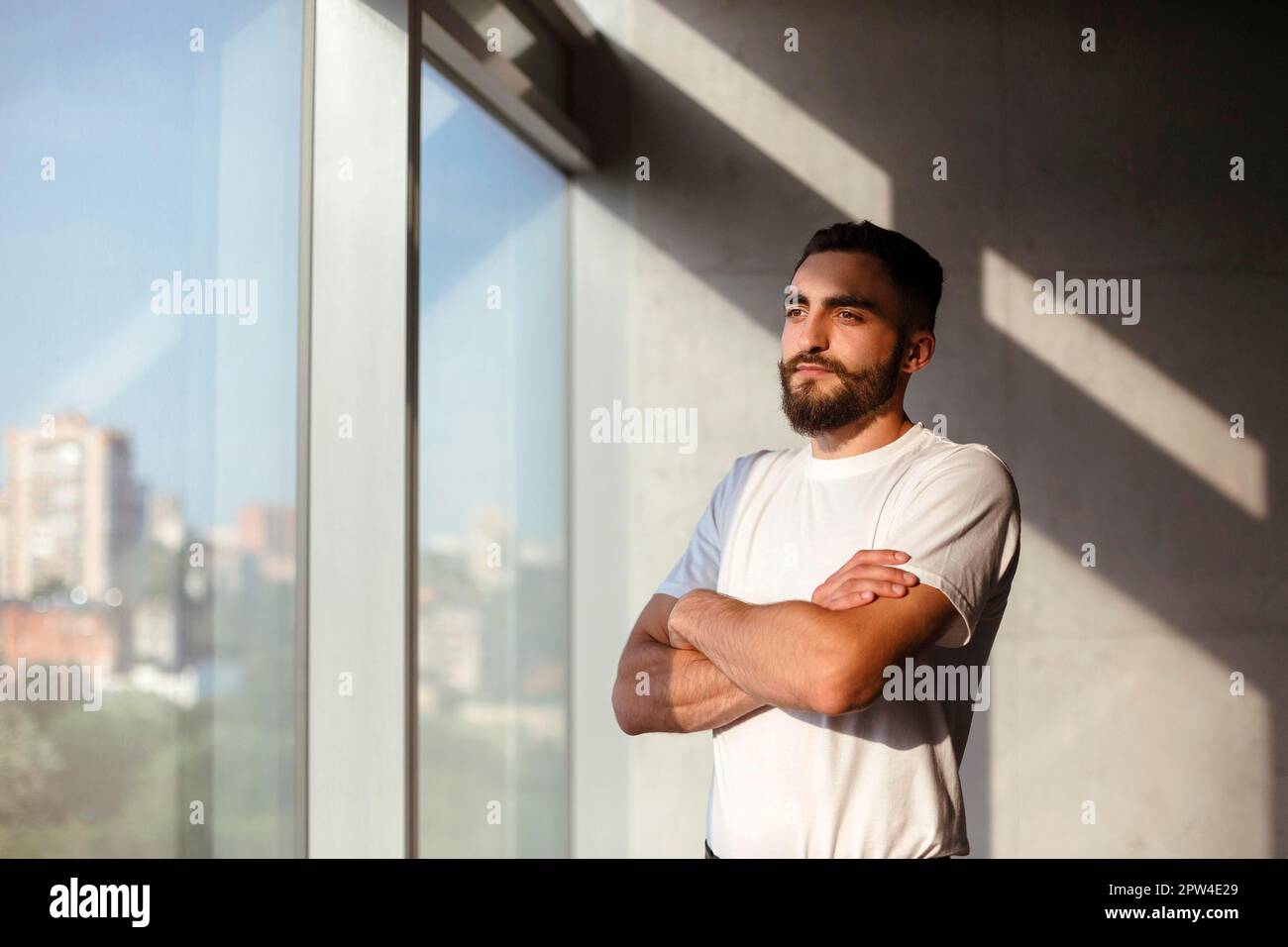 Bearded man in white t shirt looking at camera while standing near window in sunlit loft workplace Stock Photo