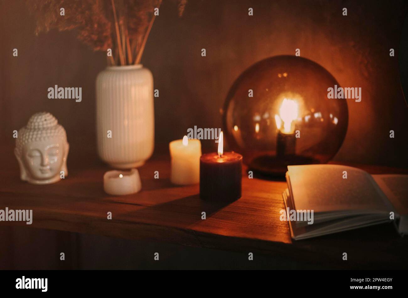 Round mirror on wooden shelf with two burning candles and small porcelain Buddha head reflected in corner of mirrored surface over concrete wall Stock Photo
