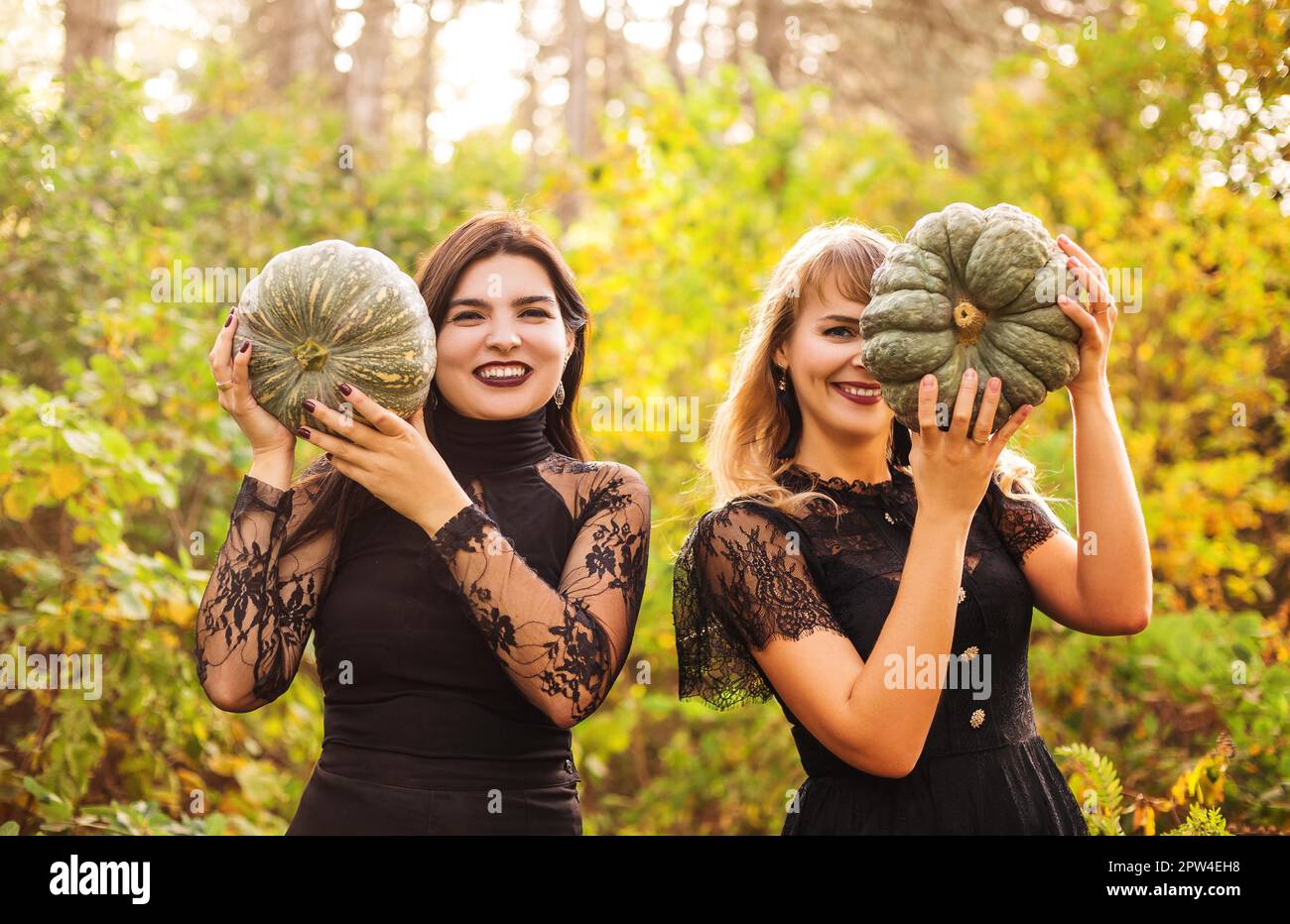 Two female friends holding scary green pumpkins in front of her face, standing isolated over autumn forest background. Fall holidays, halloween Stock Photo
