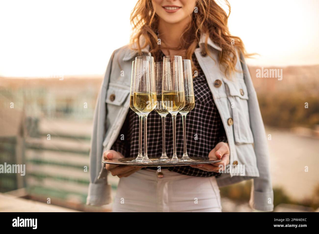 Young blonde smiling woman holding tray with champagne sparkling wine in flute glasses, standing outdoors over blurred countryside background Stock Photo