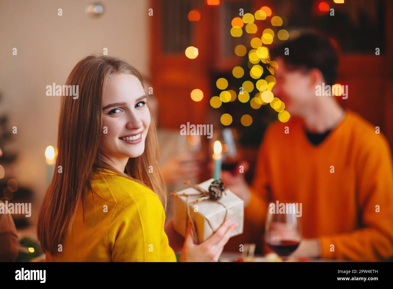 Happy smiling blond woman giving wrapped gift to friend while sitting at table and celebrating Christmas together Stock Photo