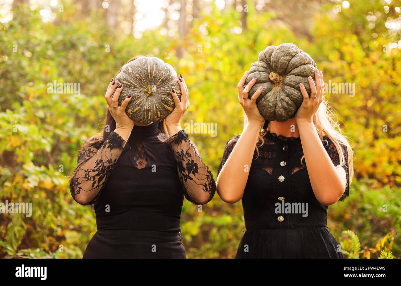Two female friends holding scary green pumpkins in front of her face, standing isolated over autumn forest background. Fall holidays, halloween Stock Photo