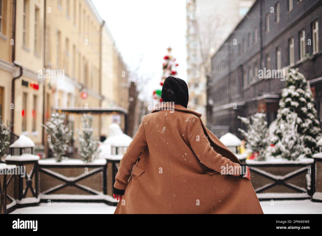 Young positive woman with outside in beige coat and black scarf on head, shivering from winter cold weather against background of trees covered with Stock Photo