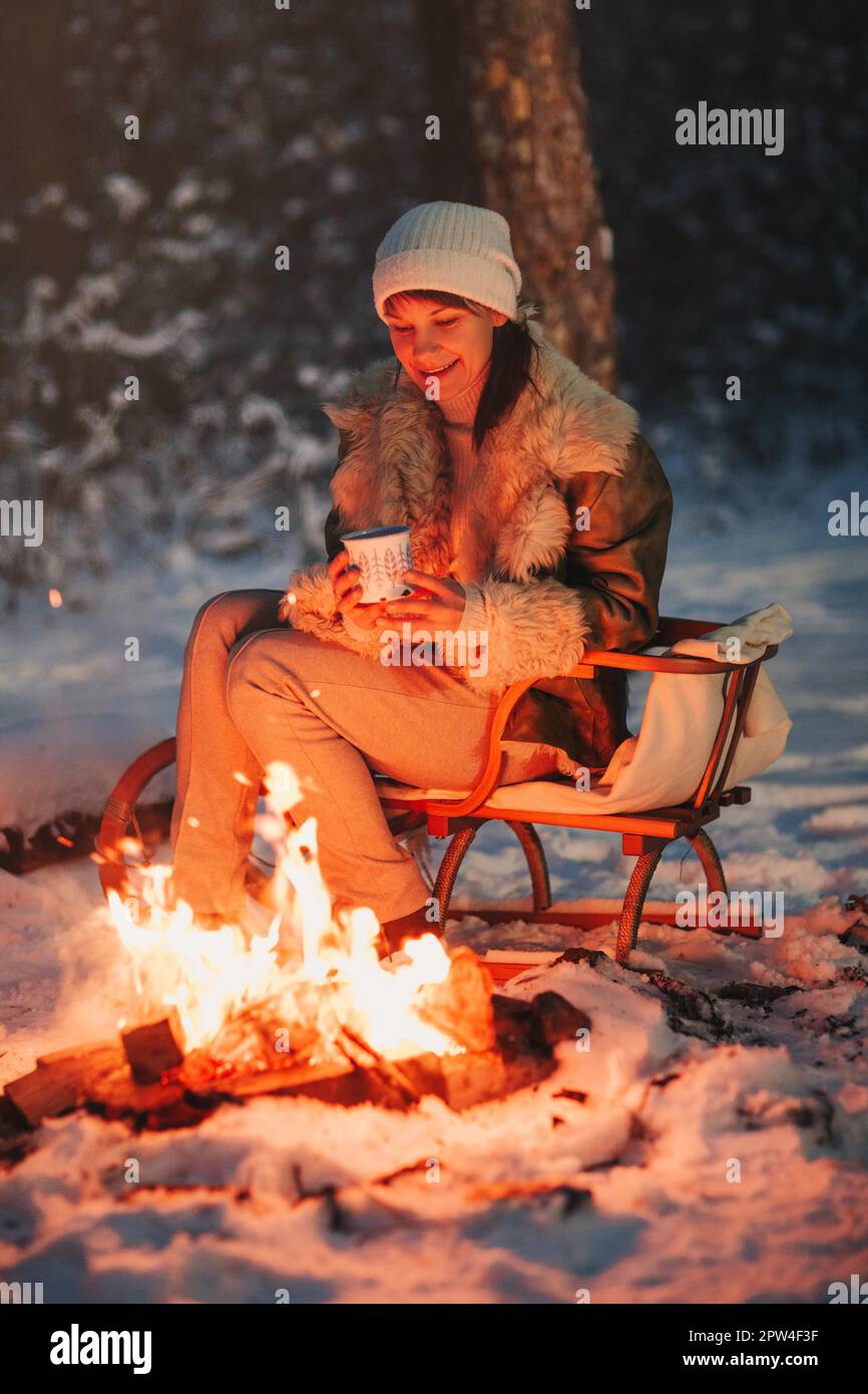 Happy young woman full of calmness and pacification in warm knitted hat and  jacket sits on wooden sled by bonfire with cup of hot tea in her hands  Stock Photo - Alamy