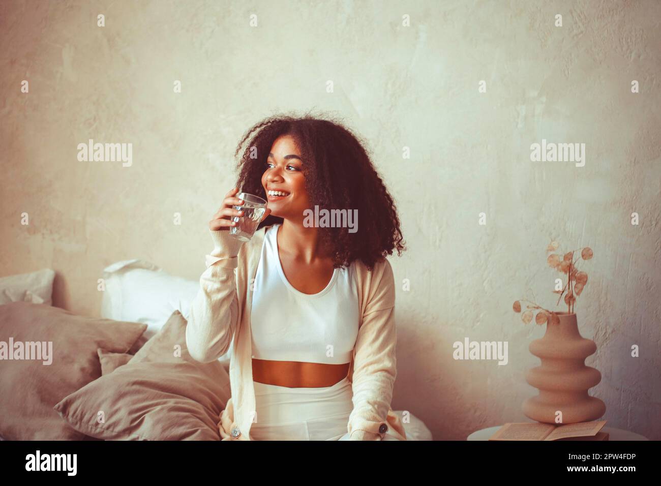 Aqua balance. Young smiling african american woman sitting at home with glass of clean mineral water, happy curly black girl enjoying healthy morning Stock Photo