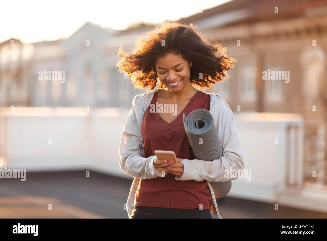 Young sporty happy african american woman in sportswear with yoga mat in hands standing on roof and using mobile phone and smiling, chatting with Stock Photo