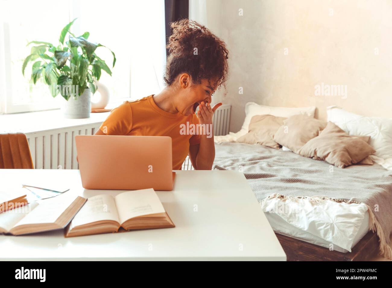 Tired flustrated African descent young girl sitting at desk in front of laptop while yawning cover mouth with hand not having interest enthusiasm for Stock Photo
