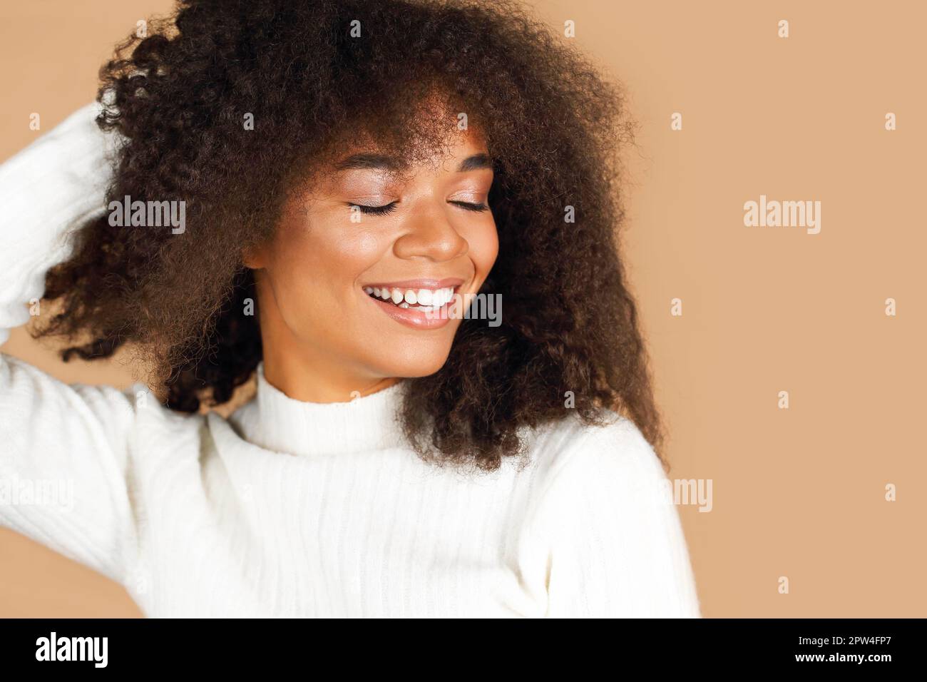 Portrait of gorgeous cheerful african american young woman with curly hair smiling while standing against beige studio background, positive black Stock Photo
