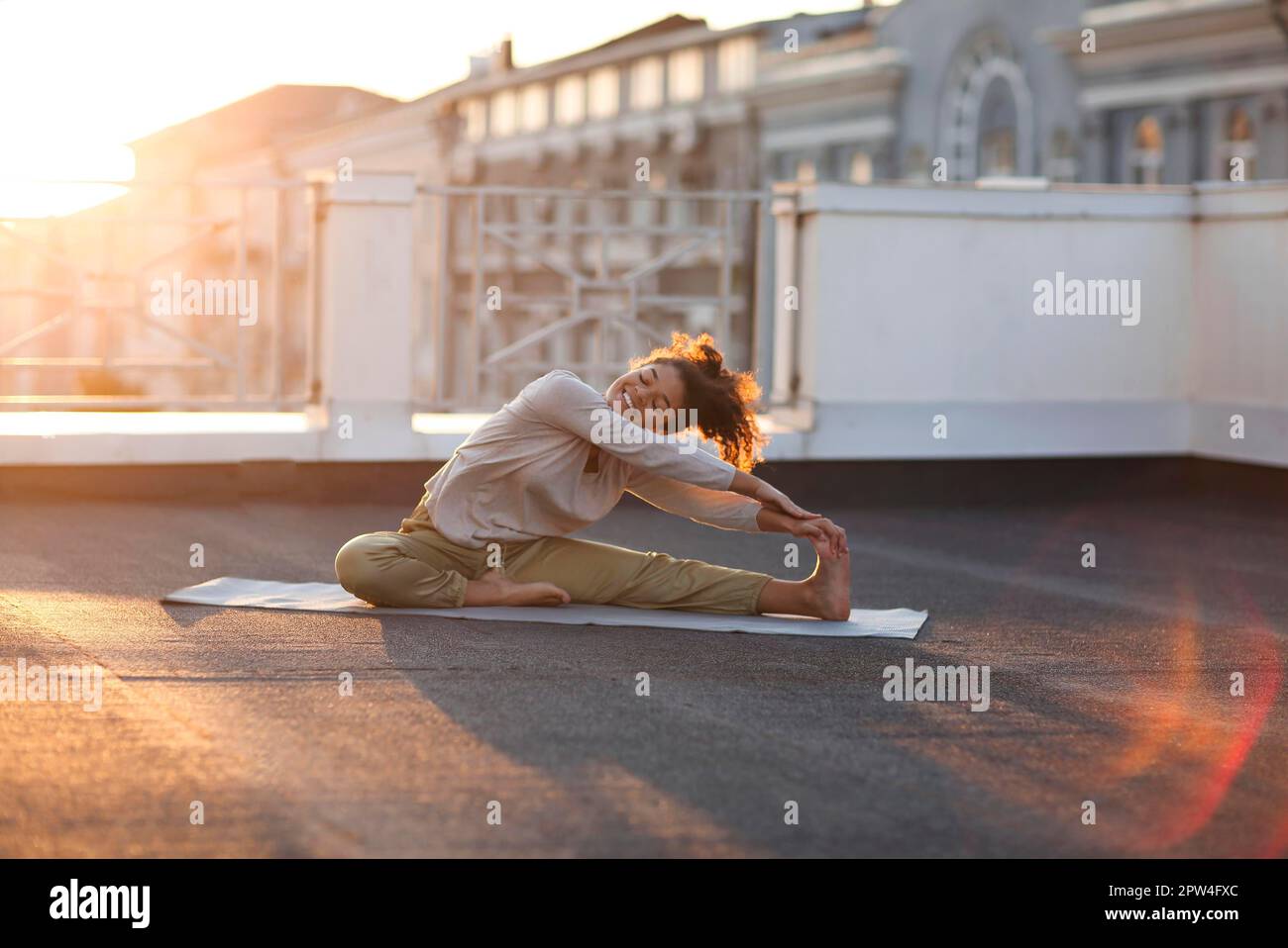 Young sporty happy mixed-race woman in sportswear doing stretching exercises while sitting on yoga mat on house roof in early morning, joyful fit Stock Photo