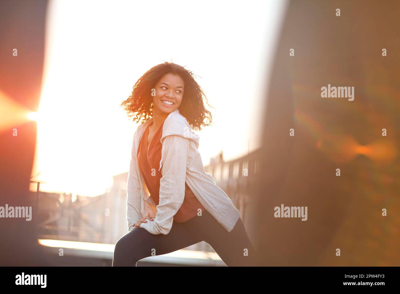 Young sporty mixed-race woman doing warming up stretching exercises and smiling while standing in morning on roof with cityscape on background Stock Photo