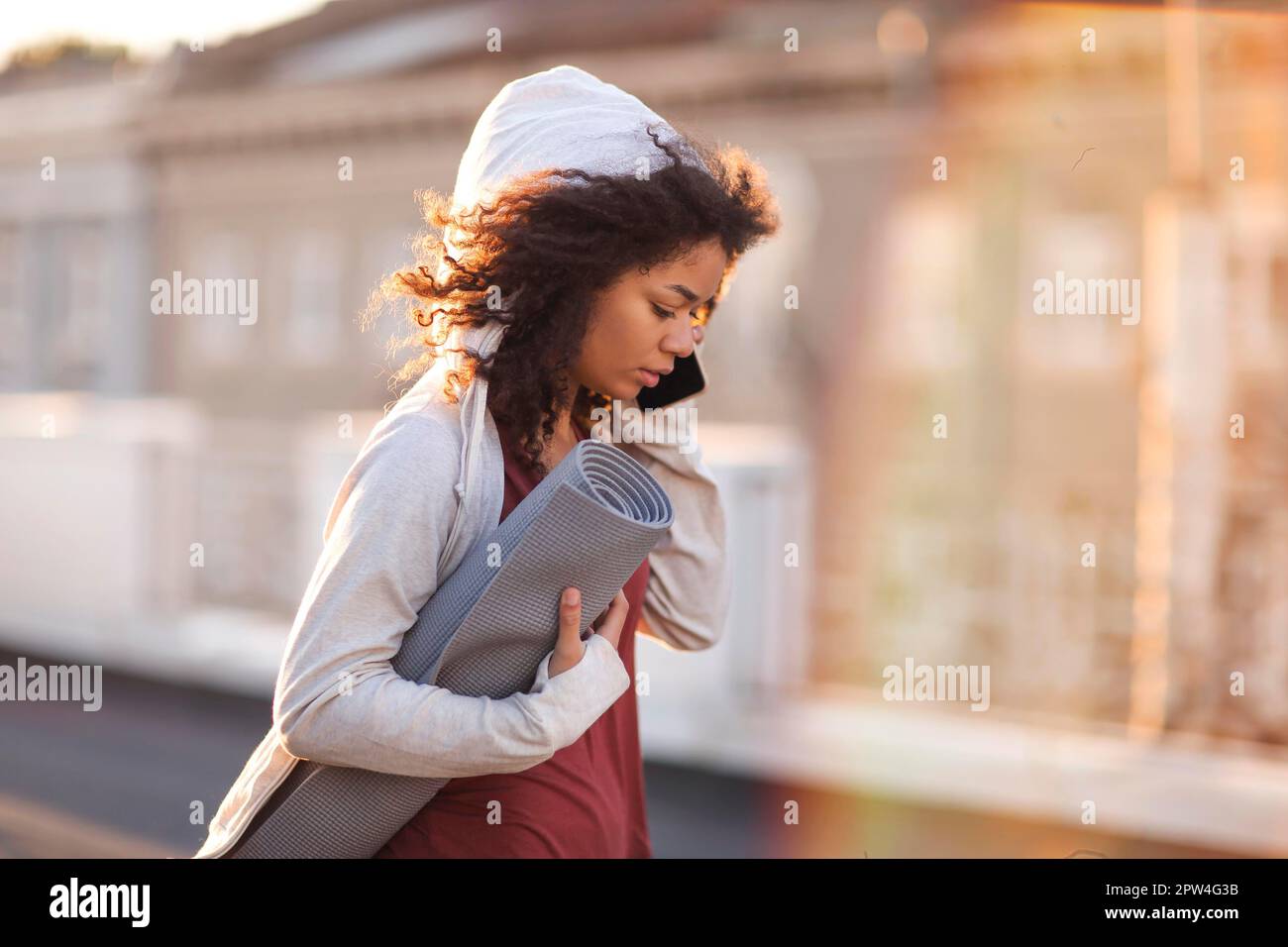 Young sporty happy african american woman in sportswear with yoga mat in hands standing on roof and using mobile phone and smiling, chatting with Stock Photo