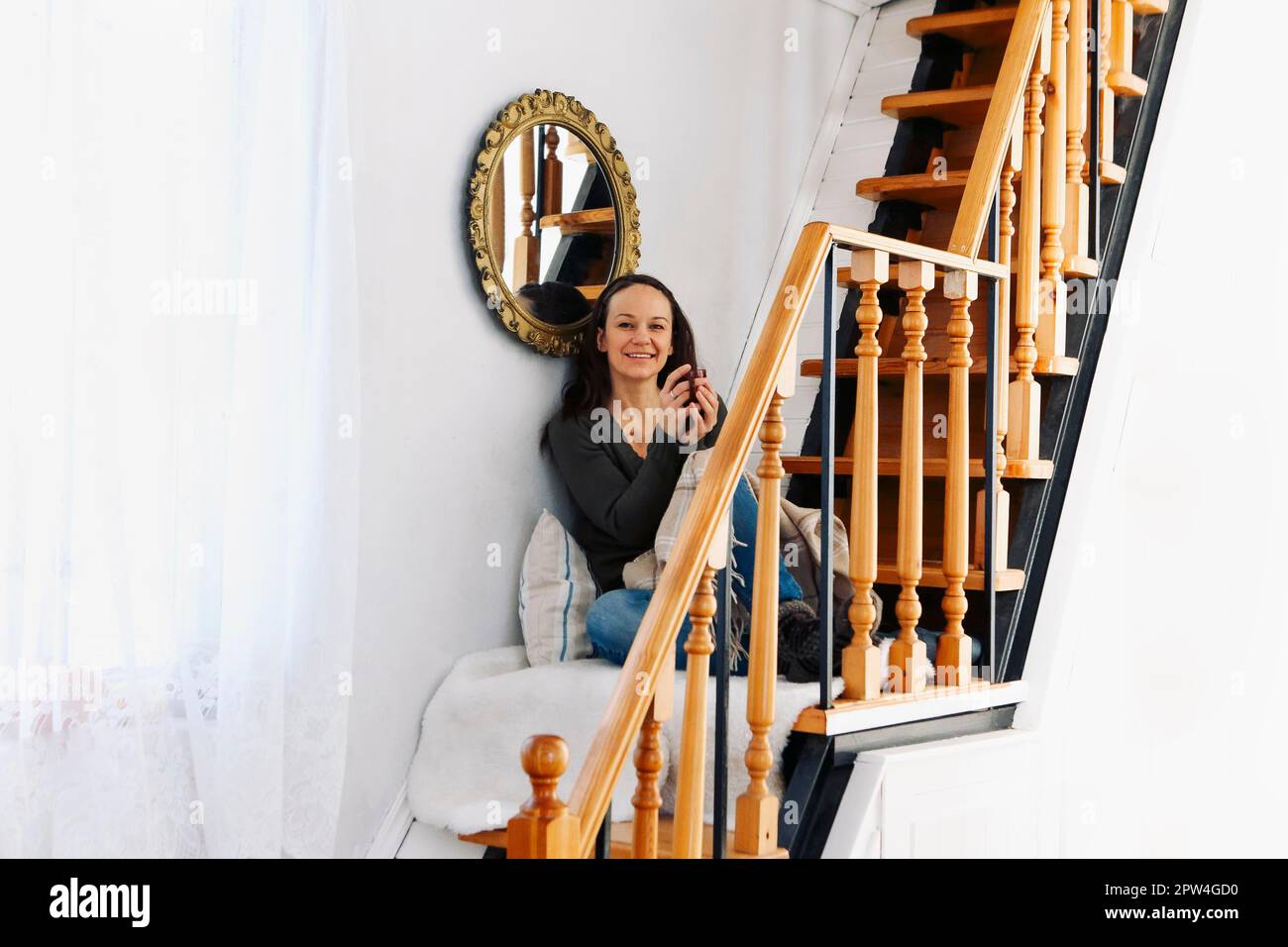 Young brunette caucasian woman sitting on a stairs at home, drinking tea and wearingwarm clothes. Positive female with open smile Stock Photo